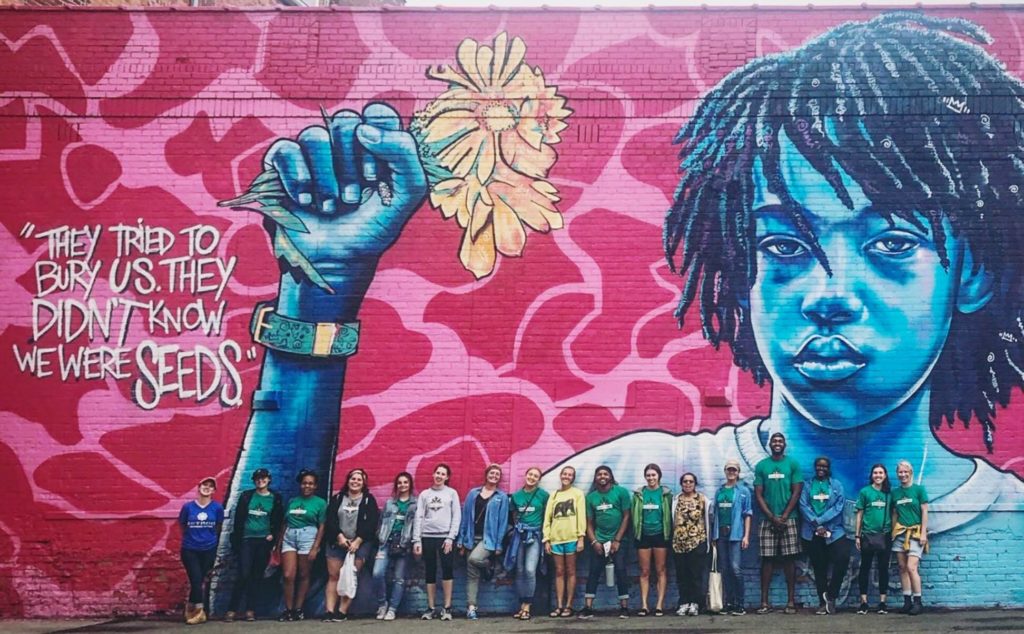 FoodCorps members standing in fronton a building with a huge painting n the side and the caption: "They tried to bury us. They didn't know we were seeds."