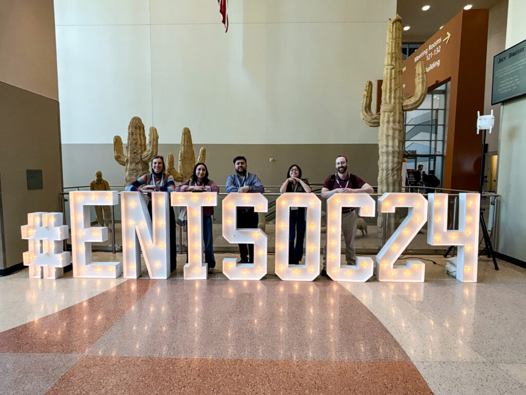 Pictured from left are Nicki Bailey ’21, Sandra Lìzarraga ’21, Marco Ponce ’19, Rina Talaba ’22, and Dan Hulbert ’09, standing behind large illuminated letters spelling out "#ENTSOC24" at a conference. They are in a modern indoor space with a cactus sculpture in the background and a digital display on the wall. The group is dressed in casual attire, wearing conference lanyards.