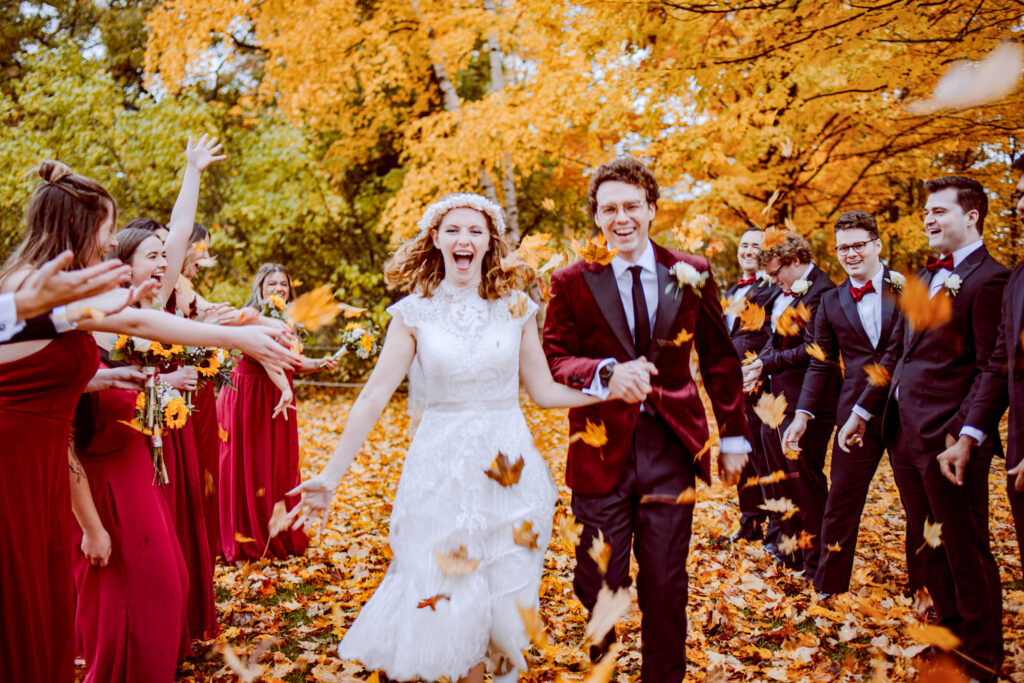 Sean Bogue ’18 and Aly Homminga ’20 are at the center of their wedding party in a vibrant autumn setting, surrounded by falling orange leaves and golden trees. Aly wears a white lace wedding dress with a floral crown, while Sean is in a maroon suit with a black tie. The bridesmaids, including Debra Farrell ’93, Lukia Artemakis ’21, Eleanor Hughes ’21, and Ynika Yuag ’21, are dressed in red gowns and hold bouquets with sunflowers. The groomsmen, including Kanwal Chowdhury ’18, Aidan Ives Johnson ’17, Graham Key ’16, Austen Scheer ’18, and Ben Homminga ’23, wear dark suits with boutonnieres. The group joyfully tosses leaves in the air