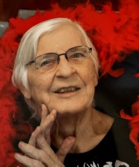 Ruthellen (Smith) McCurley '53, an older woman with white hair and glasses, smiling thoughtfully with her hand near her face, surrounded by vibrant red feathers and wearing a black top with a subtle design, against a dark background.