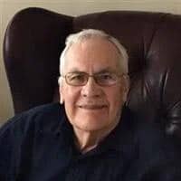 Charles True Goodsell '54, an older man with white hair and glasses, smiling while seated on a dark leather chair, wearing a dark shirt.