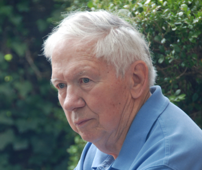 Herbert Allan Grench '54, an older man with white hair, wearing a light blue collared shirt, looking to the side with a serious expression against a background of green foliage.