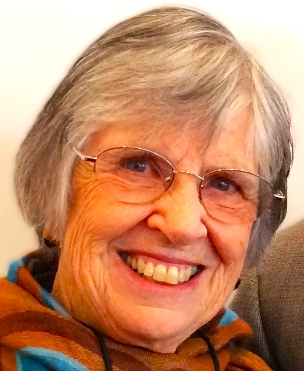 Sally (Smith) Bobertz '57, an older woman with gray hair, smiling warmly while wearing glasses and a multicolored scarf in shades of orange, blue, and brown, seated against a light background.