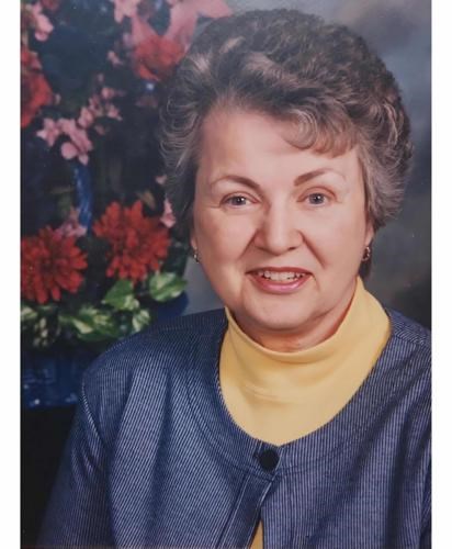 Carole (Oliver) Shick '57, an older woman with short, curly gray hair, smiling while wearing a yellow turtleneck under a blue jacket, with a vibrant bouquet of red and pink flowers in the background.