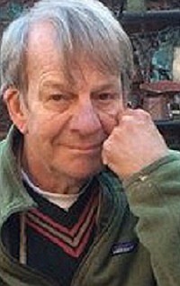 Richard Charles Newberg '58, an older man with gray hair, resting his chin on his hand while wearing a green fleece jacket over a black sweater with red and white accents, with an outdoor background featuring blurred greenery and objects.
