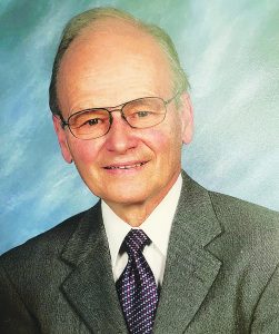 Jon M. Clapp '59, an older man with thinning gray hair and glasses, smiling while wearing a gray suit, white dress shirt, and a patterned tie in shades of purple and white, against a soft blue background.