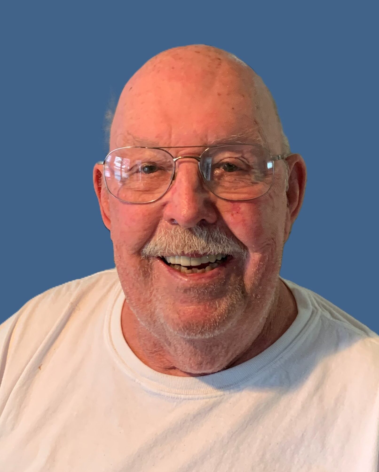 Ralph Edward Kenyon '59, an older bald man with a white mustache, smiling warmly while wearing glasses and a white T-shirt, against a solid blue background.