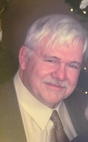 Norman Bruce Douglas '60, an older man with white hair and a mustache, smiling while wearing a white dress shirt, a patterned tie, and a dark jacket, with festive lights and greenery in the background.