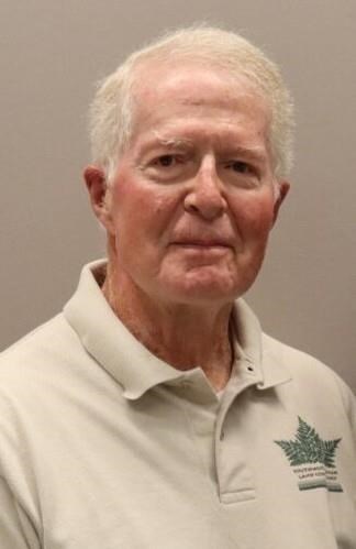 Alfred John 'Red' Gemrich '60, an older man with white hair, wearing a light beige polo shirt with a green leaf emblem, standing against a plain light brown background.