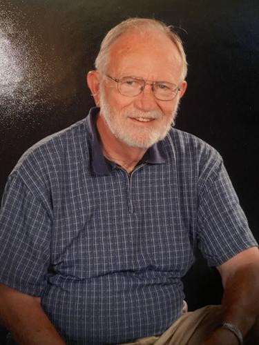 Donald S. McClure '60, an older man with thinning white hair, a white beard, and glasses, smiling while wearing a blue checkered polo shirt and beige pants, seated against a dark, speckled background.