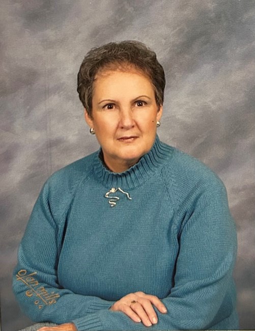 Jane Catherine (Ayers) Walsh '61, an older woman with short gray hair, wearing a teal turtleneck sweater with a small pin and the text 'Glen Mills' on the sleeve, sitting with her hands clasped against a mottled gray background.