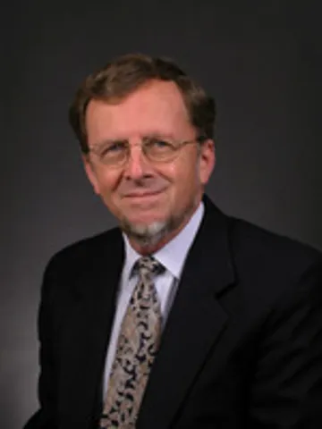 John Martin Grandin '63, a man with short brown hair and glasses, smiling while wearing a black suit, white dress shirt, and a patterned tie in shades of gray and white, against a dark gray background.