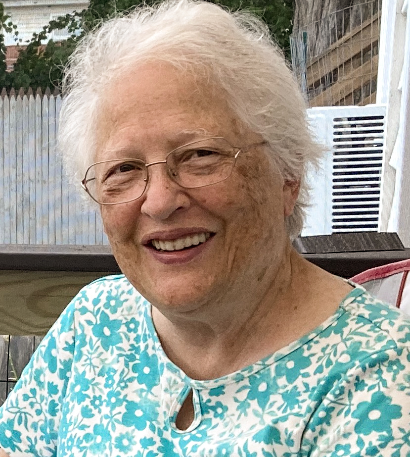 Maggie Wheeler Sadoway ’64 sits outdoors, smiling warmly. She has white hair and wears glasses and a light blue top with a white floral pattern. The background includes a white picket fence and part of a patio setting with a table and chairs.
