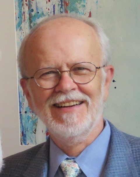 John S. Bolin '65, an older bald man with a white beard and glasses, smiling while wearing a light blue dress shirt, a floral-patterned tie, and a gray blazer, standing in front of a colorful abstract painting with splashes of blue, red, and white.