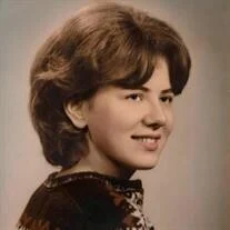 Susan Ruth (Cope) Ekstrom '68, a young woman with voluminous brown hair, smiling softly while wearing a dark top with a light-colored, patterned collar, against a neutral background.