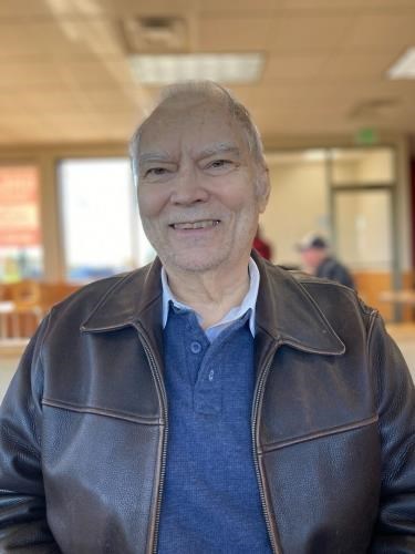 Donald Gooding Mason Jr. '68, an older man with thinning gray hair, smiling while wearing a brown leather jacket over a blue collared shirt, standing in a well-lit indoor setting with tables, chairs, and large windows in the background.