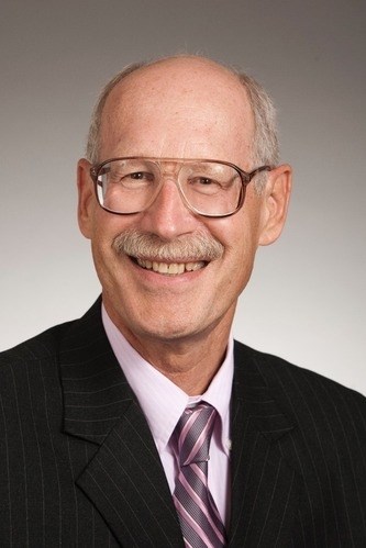 Mark B. Severs '69, an older bald man with a mustache and glasses, smiling while wearing a dark pinstripe suit, a light pink dress shirt, and a striped tie in shades of purple and white, against a gradient gray background.