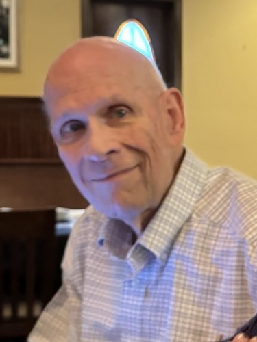 Thomas W. Thackara '69, an older bald man, smiling while wearing a light blue and white checkered shirt and a blue party hat with a star pattern, in an indoor setting with wooden furniture and a framed picture in the background.