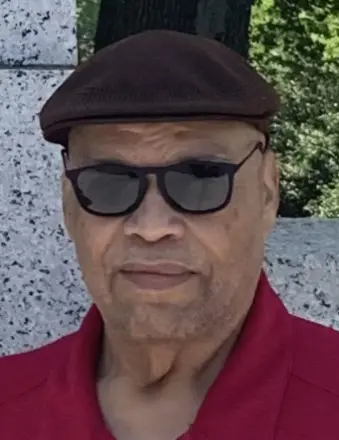 Eugene Roger Blue Jr. '72, an older man wearing a brown flat cap, dark sunglasses, and a red collared shirt, standing outdoors with a stone wall and greenery in the background.