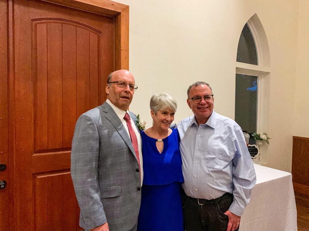 From left to right, Michael Kane ’74, Andrew Paul ’75, and Andrew's wife stand together indoors, smiling. Michael, on the left, wears a grey suit with a red tie; Andrew's wife, in the middle, is in a blue dress with a keyhole neckline; and Andrew, on the right, wears a light blue dress shirt and dark pants. They are in front of a wooden door and a table with floral decorations and a small chalkboard sign, with an arched window in the background.
