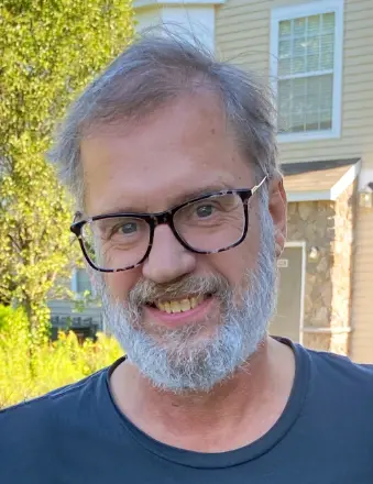 Roger Allen Gerlach '76, a man with gray hair, a white beard, and glasses, smiling while wearing a dark blue shirt, standing outdoors in front of a house with stone and beige siding, surrounded by green foliage.
