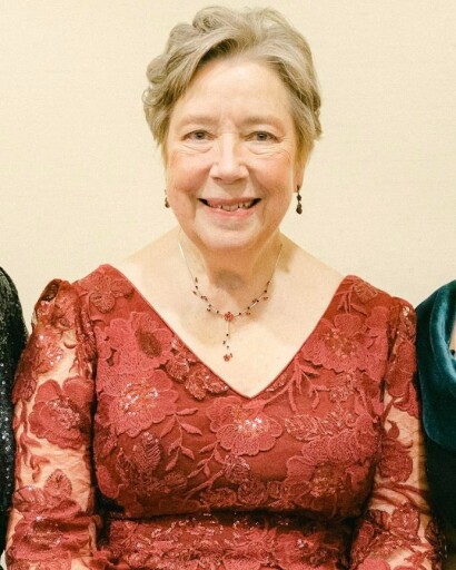 Susan M. Smoley '77, an older woman with short gray hair, smiling while wearing a deep red lace dress with a V-neckline, a matching necklace, and drop earrings, seated against a light background.