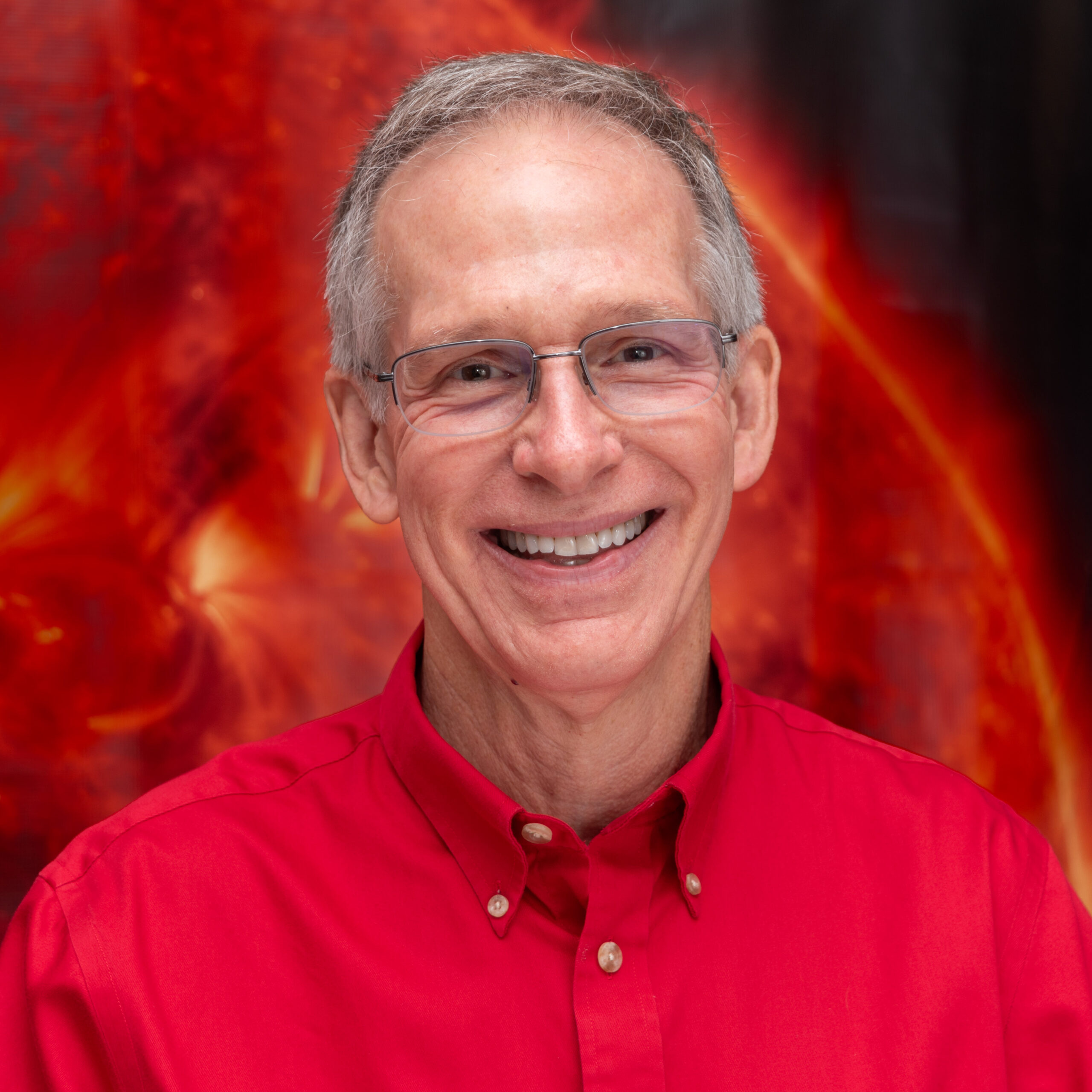 James Klimchuk ’79 stands against a vibrant, fiery red and orange background that resembles an abstract cosmic scene. He wears a bright red button-up shirt with a collar, and his hair is grey.