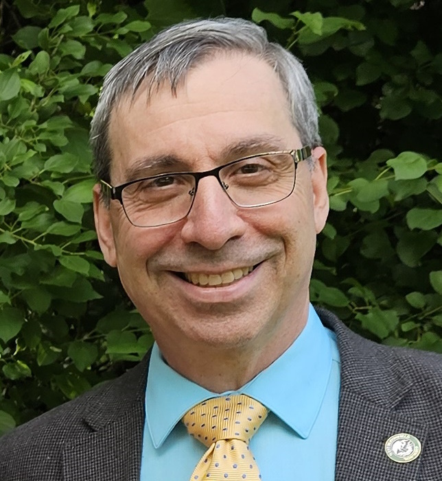 Jonathan Warshay ’82 stands outdoors with a backdrop of lush green foliage. He has grey hair and wears a dark blazer over a light blue dress shirt, paired with a yellow tie featuring a blue polka dot pattern. A small pin is visible on his blazer lapel.