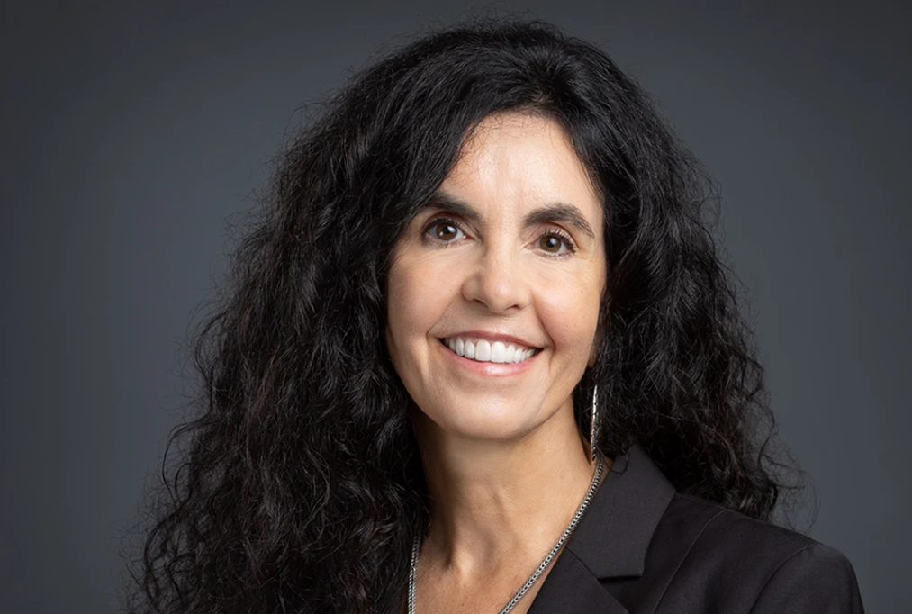 Cristin Reid ’90 poses for a portrait against a dark grey background. She has long, curly black hair and wears a black blazer. A silver necklace is visible around her neck, and she has a pair of dangling earrings.