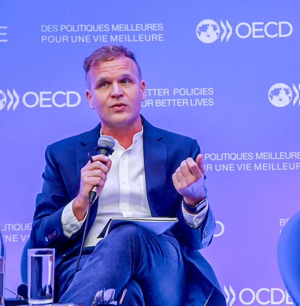 Michael L. Bąk ’96 is seated on a blue chair during an event, speaking into a microphone. He wears a navy suit jacket over a white dress shirt and jeans, holding a mic one hand while gesturing with the other. The background features a blue backdrop with the OECD logo and the text "DES POLITIQUES MEILLEURES POUR UNE VIE MEILLEURE" (Better Policies for Better Lives). A glass of water and some papers are on the table beside him.