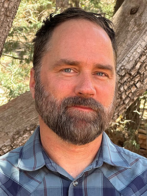 Christopher Lyons ’96 stands outdoors in a natural setting with trees and greenery in the background. He has a beard and short hair, and is wearing a blue and white plaid button-up shirt.
