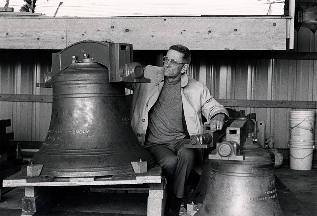 Professor Emeritus T. Jefferson Smith sits in a workshop surrounded by large metal bells. He wears glasses, a light coat, and a sweater, and is positioned near a bell on a wooden pallet, with another bell and workshop materials like buckets in the background. The black-and-white photo has a vintage feel.