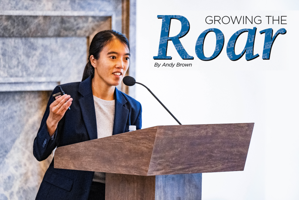 Amanda Moss ’19 stands at a wooden podium, speaking into a microphone during a presentation. She wears a dark blazer over a light top and holds a small device in her hand, gesturing as she speaks. The background is a light-colored wall. Overlaid text in the right corner in blue reads "GROWING THE ROAR" with "By Andy Brown" in smaller black text below.