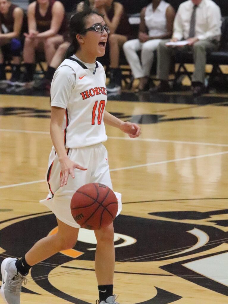 Amanda Moss ’19 wearing a white jersey with "HORNETS" and the number 10 is dribbling a ball on the court. She appears to be in motion, with a focused expression, during a game. Opposing team benches are visible in the background.