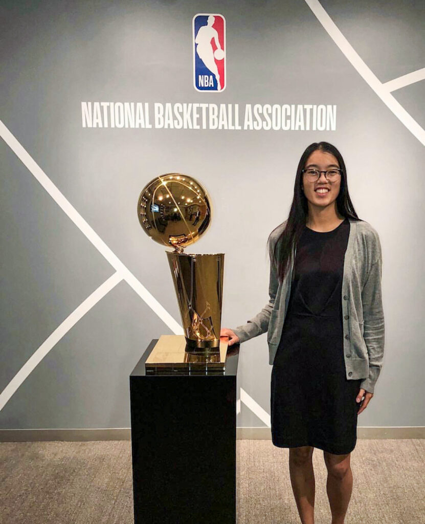 Amanda Moss ’19 stands smiling next to a shiny NBA championship trophy on a black pedestal. She wears a black dress with a grey cardigan. The background features the NBA logo and the words "NATIONAL BASKETBALL ASSOCIATION" on a grey wall with white geometric lines.