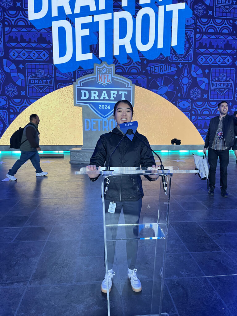 Amanda Moss ’19 stands at a clear podium with an NFL microphone, at the 2024 NFL Draft in Detroit. She wears a black jacket, light blue pants, and white sneakers. The backdrop features the NFL Draft logo, the word "DETROIT," and a blue and gold design with "2024" repeated. A few people are visible in the background, including one with a backpack and another holding a white bag