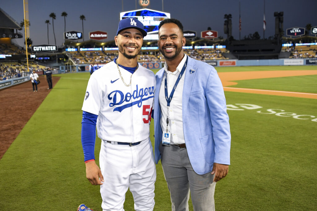 Jordan Wiley ’19 and Mookie Betts stand together on the field at Dodger Stadium at night. Mookie Betts, on the left, wears a white Los Angeles Dodgers uniform with the number 50 and a blue cap, while Jordan Wiley ’19, on the right, is dressed in a light blue blazer over a white shirt, with a Dodgers lanyard around his neck. The stadium is well-lit, with a scoreboard, palm trees, and advertisements in the background.