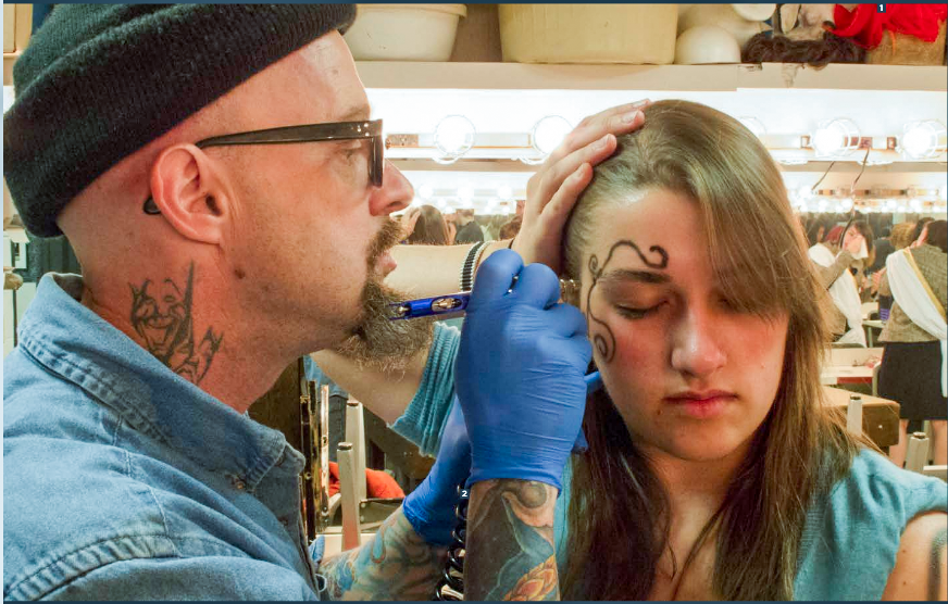 Jon A. Reeves, a bald man with a beard, wearing glasses, a black beanie, and a denim jacket, applying temporary tattoos to a young woman's forehead with blue-gloved hands in a busy room with mirrors, lights, and various items on shelves in the background.