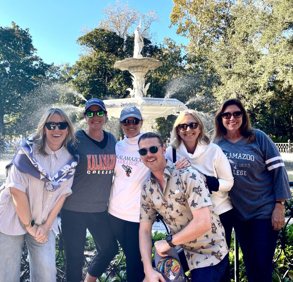 From left to right, Meredith (Robertson) Eaton, Christy (Kortryk) DeGrendel, Allison Pudduck, Anne (Herrnstein) Morris, and Erin (O’Brien) Whaley pose together in front of a white fountain, set against a backdrop of trees and a clear blue sky. Meredith wears a striped shirt with a scarf, Christy sports a grey Kalamazoo College sweatshirt, Allison is in a white Kalamazoo College hoodie and cap, Anne wears a white sweatshirt, and Erin is in a blue Kalamazoo College shirt. A man in a patterned shirt stands in the center, smiling with sunglasses on.