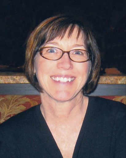 Susanne Marie Gibson, a woman with short brown hair and glasses, smiling while wearing a black top, seated against a dark background with a patterned red and beige cushion.