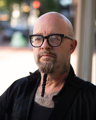 Jon A. Reeves, a bald man with a gray beard and glasses, wearing a black shirt and jacket, with a braided ponytail hanging from his chin and visible tattoos on his neck, standing outdoors near a window with a blurred street scene in the background.