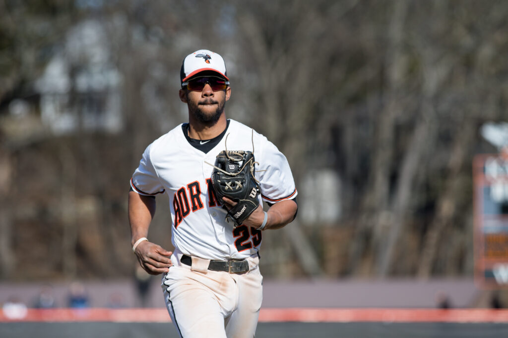 Jordan Wiley ’19 is on a baseball field, wearing a white uniform with "Hornets" written in orange across the chest and the number 25 on the lower left side of his jersey. He holds a black Wilson glove in his left hand and wears a white cap with an orange and black logo. The background shows a blurred outdoor setting with bare trees