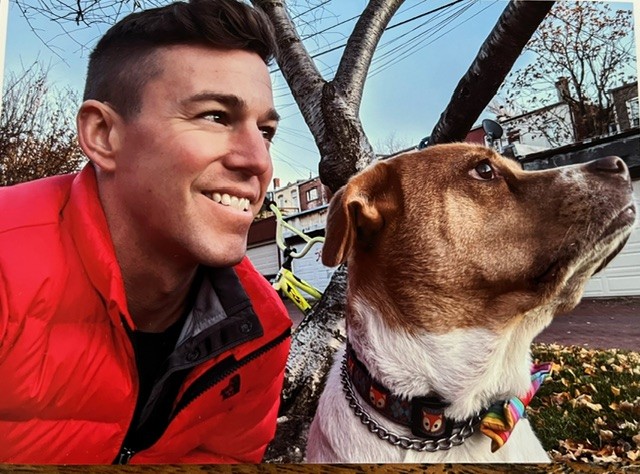 A photo of Todd Jack Robosan ’06, wearing a red jacket, smiling outdoors next to his dog Rye. The dog, with brown and white fur, wears a colorful collar and looking up to the right. They are positioned near a tree with autumn leaves and a residential background with houses and a garage door.