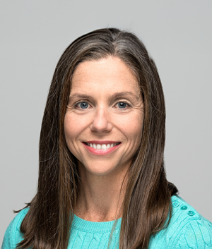 A portrait of Diana Daly ’01, with long, straight, graying brown hair, wearing a teal sweater with buttons on the shoulder. The background is a plain, neutral gray.