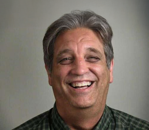 Kenneth Lyle Hill '76, a man with gray hair, smiling while wearing a green and white checkered collared shirt, against a plain light gray background.
