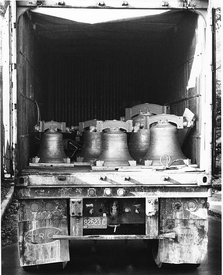 A black-and-white photo shows the open back of a truck with a license plate reading "82523 C." Inside, eight large English tower bells are secured, ready for installation. The truck's rear door is open, revealing the bells' against the dark interior.