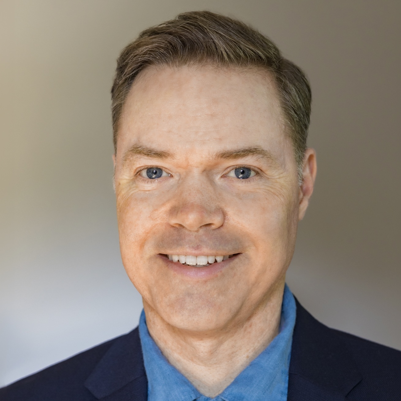 A professional headshot portrait of Carl Roose, a man with neatly combed light brown hair, smiling warmly with crinkled eyes. He wears a navy blue suit jacket over a light blue collared shirt against a soft beige background.