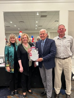 Four individuals stand together in a conference room with a mirrored wall and ceiling lights, holding a framed certificate. From left, a woman with blonde hair wears a green floral jacket and black outfit, smiling softly. Next to her, another woman with blonde hair in a black top holds the certificate, smiling warmly. In the center, a man in a blue suit and tie stands proudly, also holding the certificate. On the right, a man with short gray hair wears a checkered shirt and beige pants, smiling gently. Red, white, and blue balloons and a table with awards are visible in the background, suggesting a celebratory event.