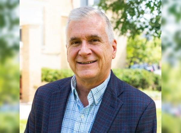 A portrait of Kevin R. Howley ’81, a man with short gray hair, smiling warmly. He is dressed in a blue checkered shirt and a dark plaid blazer. The background features a blurred outdoor setting with green trees, shrubs, and a beige building, suggesting a campus or garden environment on a sunny day.