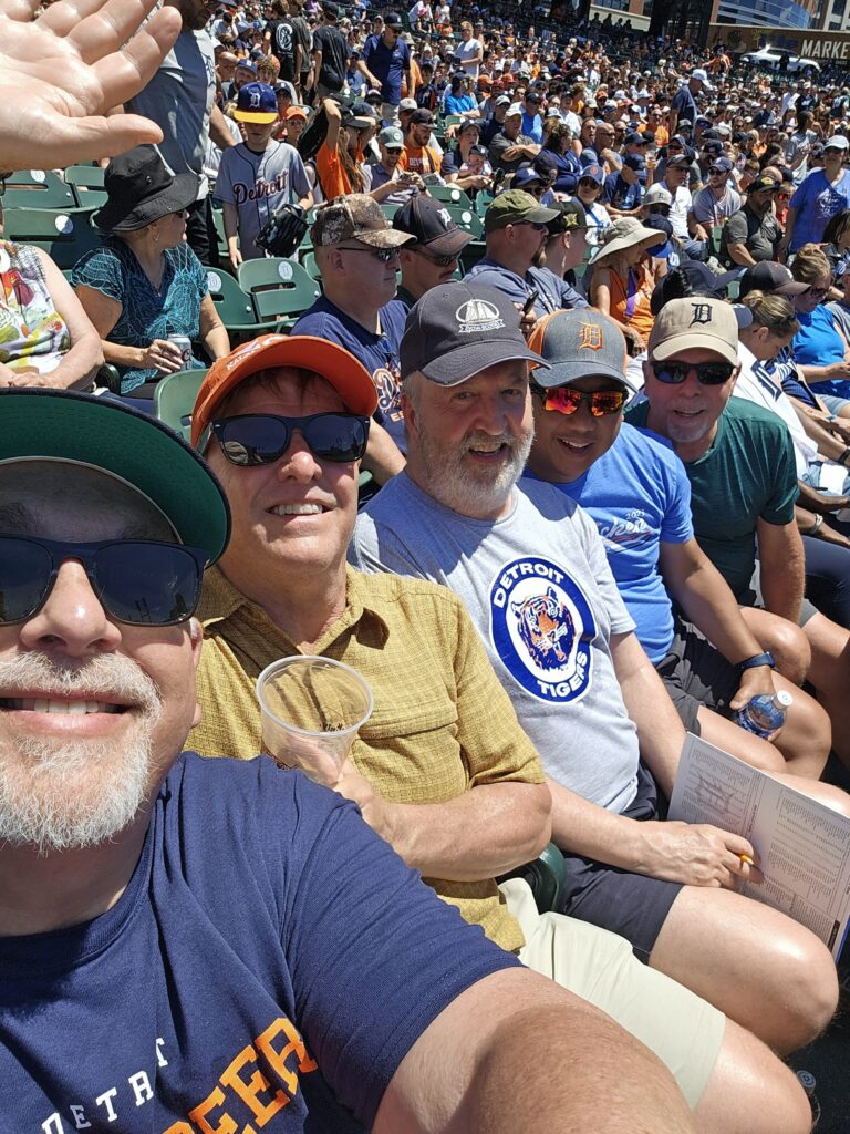A group of five people sits in a crowded baseball stadium with a large audience in the background, enjoying a sunny day. The person taking the selfie, on the far left, wears a green cap and a dark blue "Detroit Tigers" shirt, smiling with a plastic cup in hand. Next to them, a man in an orange cap and yellow shirt grins widely. In the center, a man with a white beard wears a gray "Detroit Tigers" shirt and holds a scorecard. To his right, a man in a blue shirt smiles, and on the far right, a man in a green shirt holds a water bottle. The crowd behind them wears a mix of team colors, with some in Detroit Tigers gear, and the stadium seats are filled with green chairs.